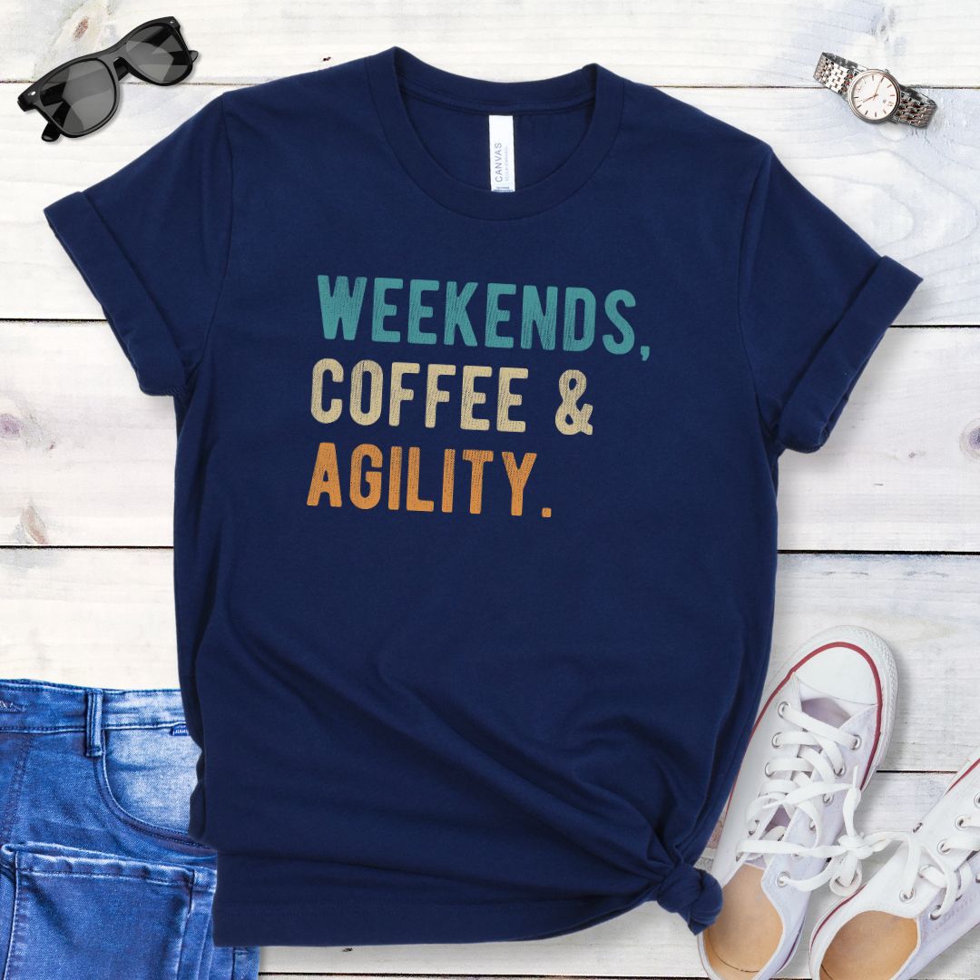 Navy blue dog sports t-shirt with 'Weekends, Coffee & Agility' in bold, colorful text, presented on a wooden surface with jeans and shoes.