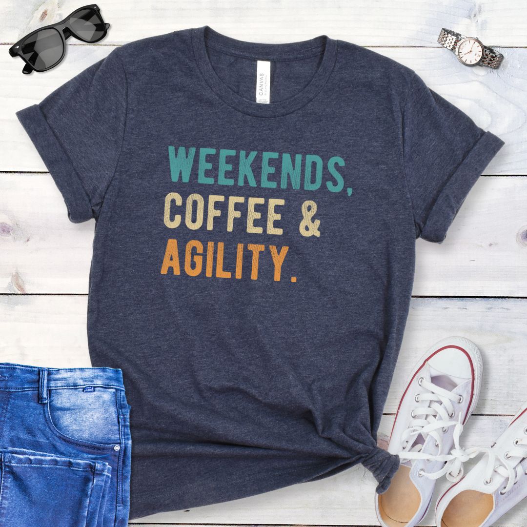 Heather navy dog sports t-shirt with 'Weekends, Coffee & Agility' in bold, colorful text, presented on a wooden surface with jeans and shoes.