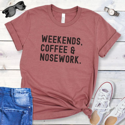 Mauve t-shirt for dog scent work trainers with 'Weekends, Coffee & Nosework' text on a wooden surface with jeans and shoes.