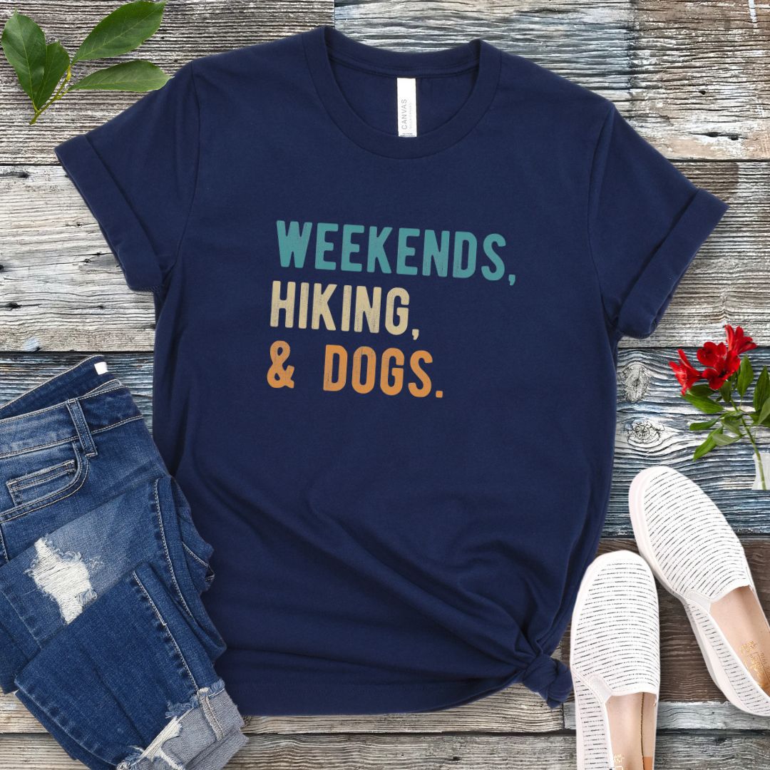 A navy unisex dog lover T-shirt with the text 'Weekends, hiking, & dogs.' printed in colorful letters, displayed on a wooden surface alongside denim jeans and white shoes.