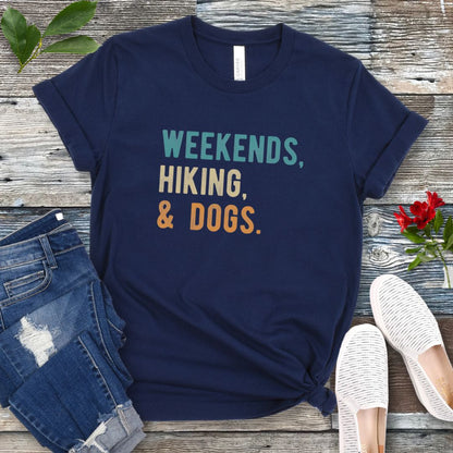 A navy unisex dog lover T-shirt with the text 'Weekends, hiking, & dogs.' printed in colorful letters, displayed on a wooden surface alongside denim jeans and white shoes.