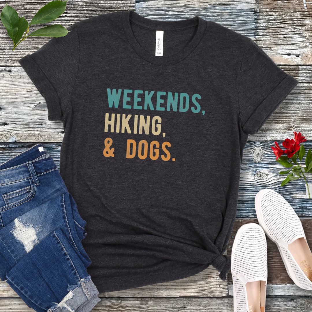 A dark grey unisex dog lover T-shirt with the text 'Weekends, hiking, & dogs.' printed in colorful letters, displayed on a wooden surface alongside denim jeans and white shoes.