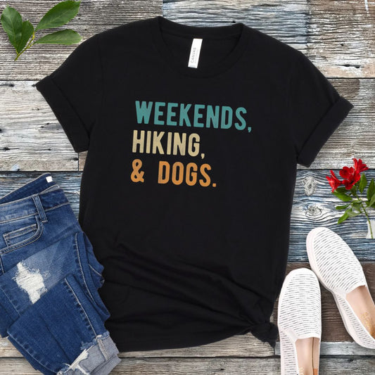 A black unisex dog lover T-shirt with the text 'Weekends, hiking, & dogs.' printed in colorful letters, displayed on a wooden surface alongside denim jeans and white shoes.