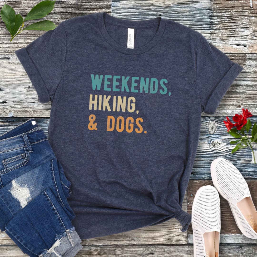 A heather navy unisex dog lover T-shirt with the text 'Weekends, hiking, & dogs.' printed in colorful letters, displayed on a wooden surface alongside denim jeans and white shoes.