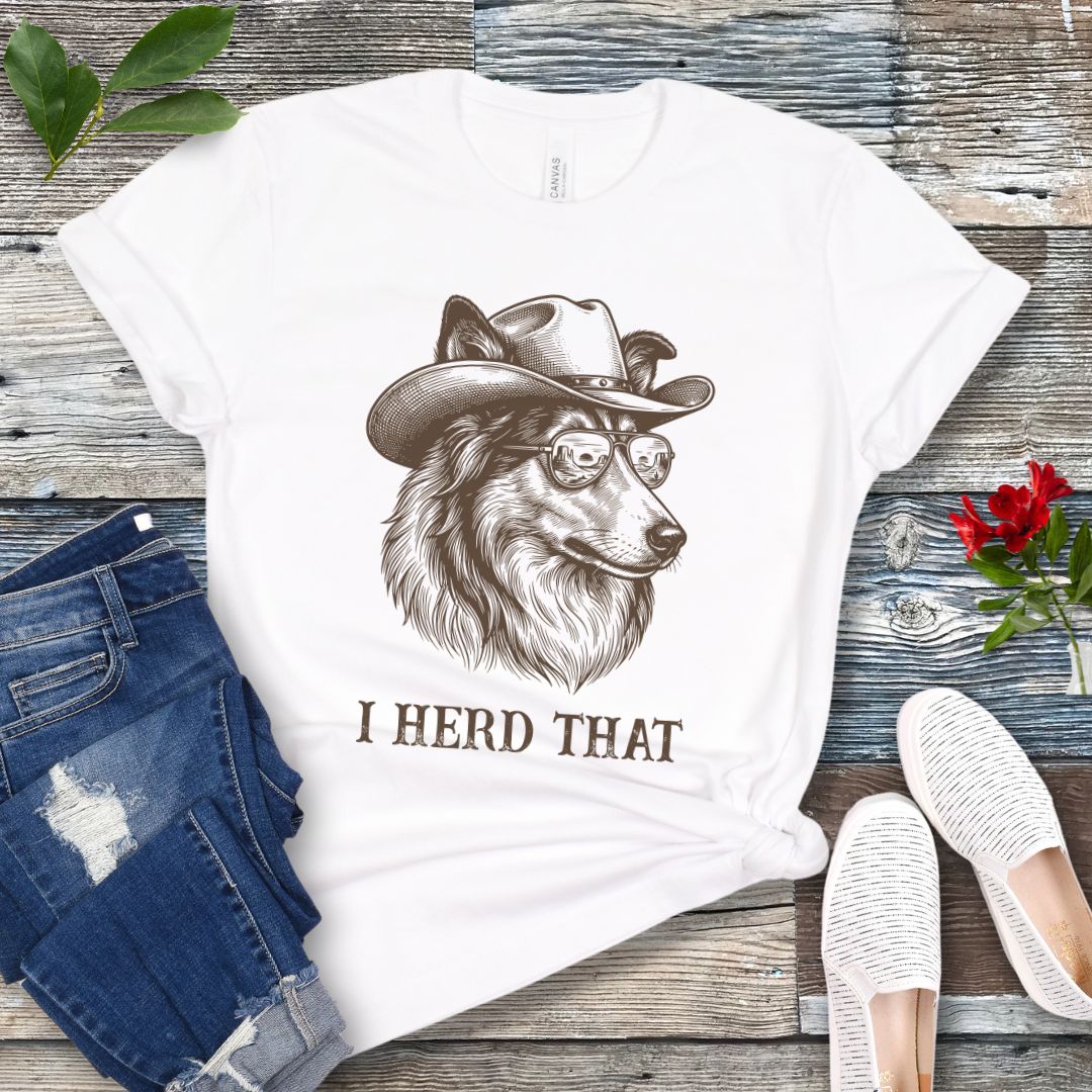 White t-shirt for dog lovers with a Rough Collie wearing a cowboy hat graphic and humorous 'I Herd That' text, placed on a wooden surface with jeans and shoes.