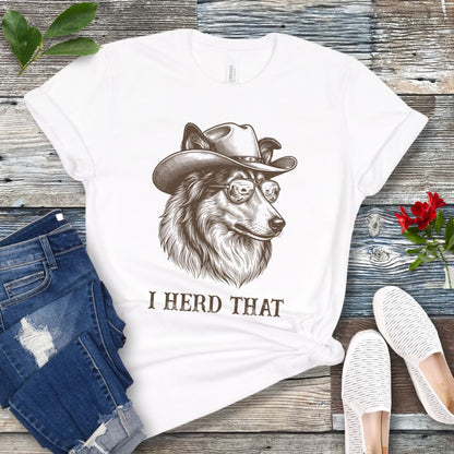 White t-shirt for dog lovers with a Rough Collie wearing a cowboy hat graphic and humorous 'I Herd That' text, placed on a wooden surface with jeans and shoes.
