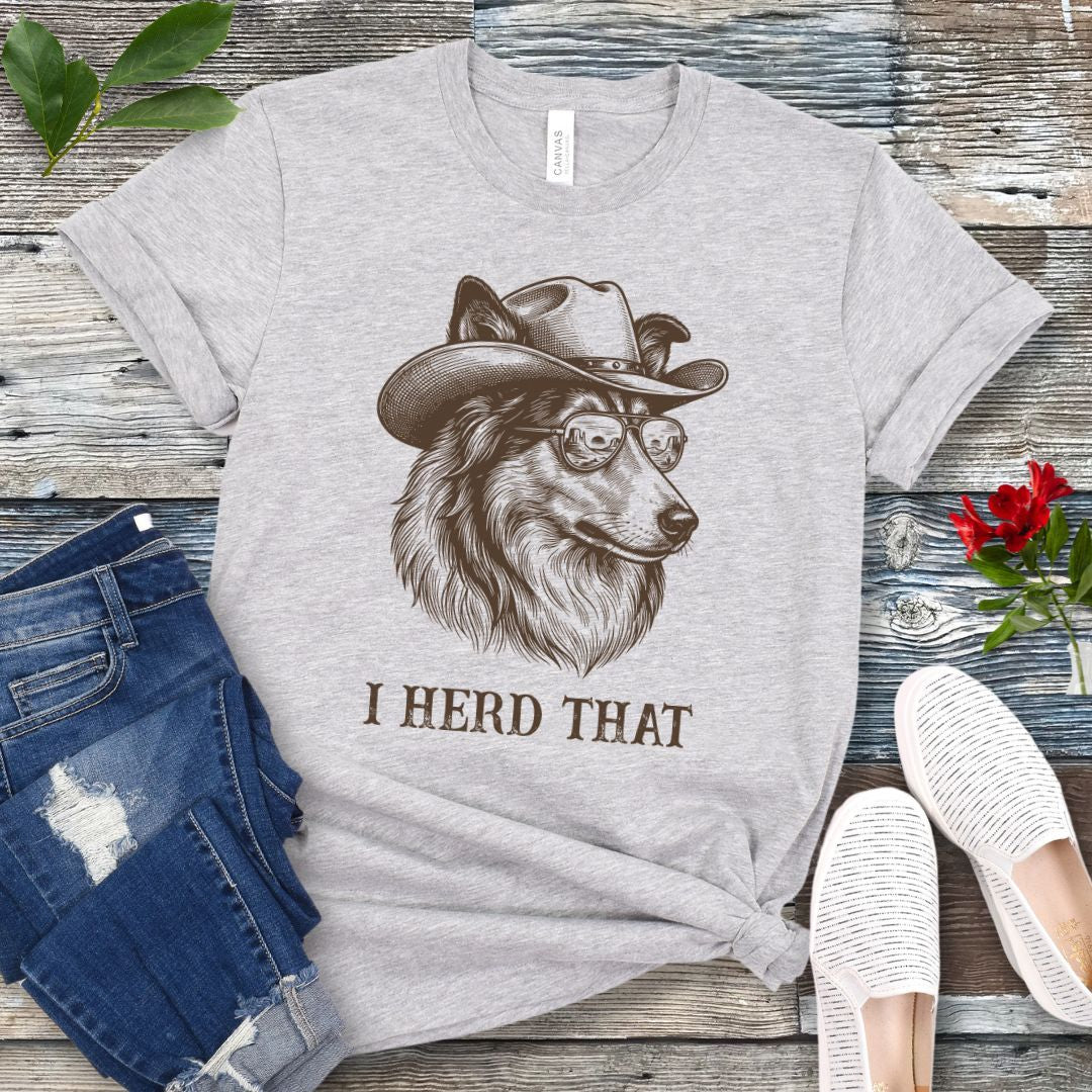 Grey t-shirt for dog lovers with a Rough Collie wearing a cowboy hat graphic and humorous 'I Herd That' text, placed on a wooden surface with jeans and shoes.