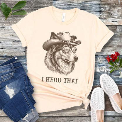 Beige t-shirt for dog lovers with a Rough Collie wearing a cowboy hat graphic and humorous 'I Herd That' text, placed on a wooden surface with jeans and shoes.