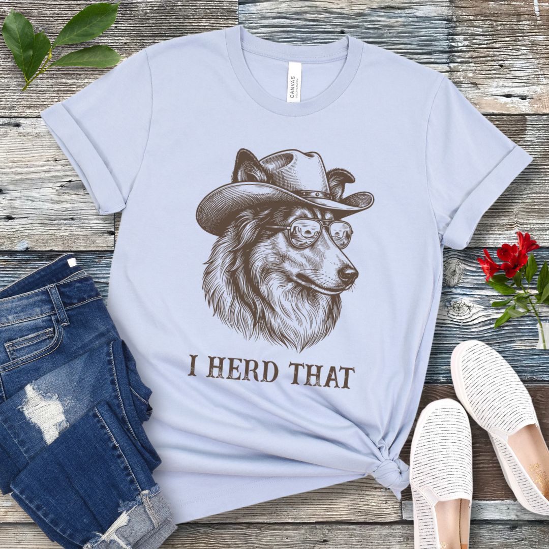 Light blue t-shirt for dog lovers with a Rough Collie wearing a cowboy hat graphic and humorous 'I Herd That' text, placed on a wooden surface with jeans and shoes.
