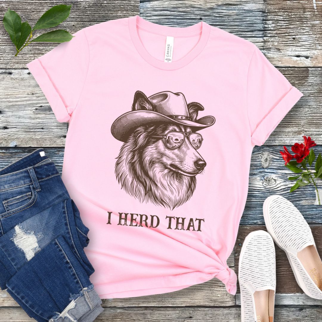 Pink t-shirt for dog lovers with a Rough Collie wearing a cowboy hat graphic and humorous 'I Herd That' text, placed on a wooden surface with jeans and shoes.