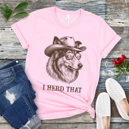 Pink t-shirt for dog lovers with a Rough Collie wearing a cowboy hat graphic and humorous 'I Herd That' text, placed on a wooden surface with jeans and shoes.