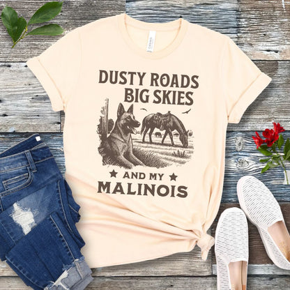 Beige t-shirt with graphic of Malinois in a rural scene and quote on a wooden surface with jeans and shoes.