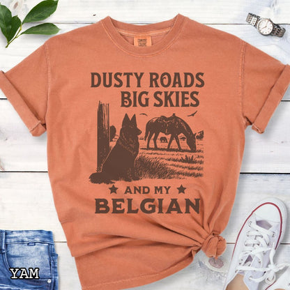 Orange Comfort Colors t-shirt featuring a Western-style design with grazing horse, rustic fence, and a Belgian Sheepdog illustration; text reads “Dusty Roads, Big Skies, and My Belgian.”