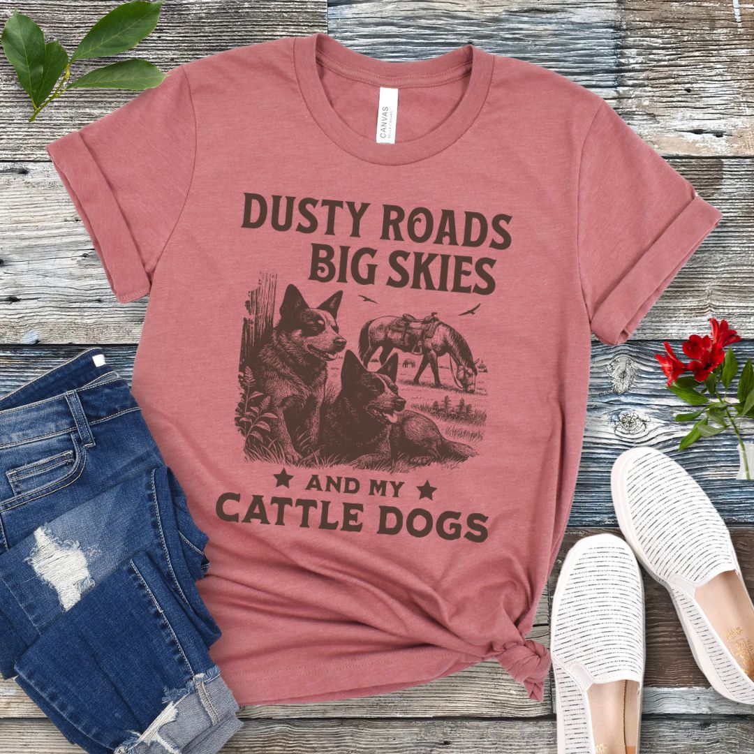Mauve western-aesthetic t-shirt with graphic of dogs and text 'Dusty roads, big skies, and my cattle dogs' on a wooden surface with jeans and shoes.