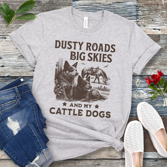 Gray western-aesthetic t-shirt with graphic of dogs and text 'Dusty roads, big skies, and my cattle dogs' on a wooden surface with jeans and shoes.