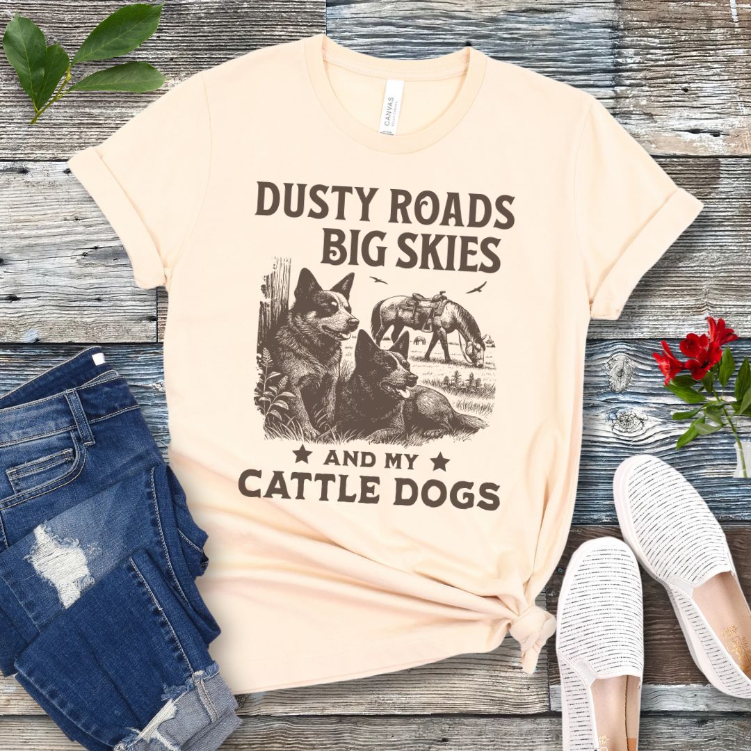 Beige western-aesthetic t-shirt with graphic of dogs and text 'Dusty roads, big skies, and my cattle dogs' on a wooden surface with jeans and shoes.
