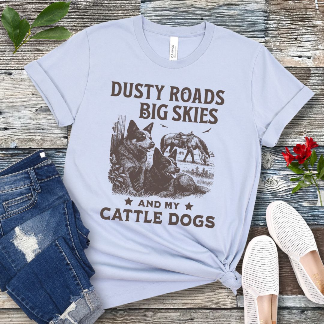 Light blue western-aesthetic t-shirt with graphic of dogs and text 'Dusty roads, big skies, and my cattle dogs' on a wooden surface with jeans and shoes.