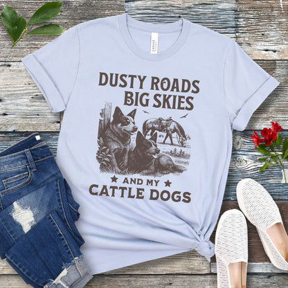 Light blue western-aesthetic t-shirt with graphic of dogs and text 'Dusty roads, big skies, and my cattle dogs' on a wooden surface with jeans and shoes.