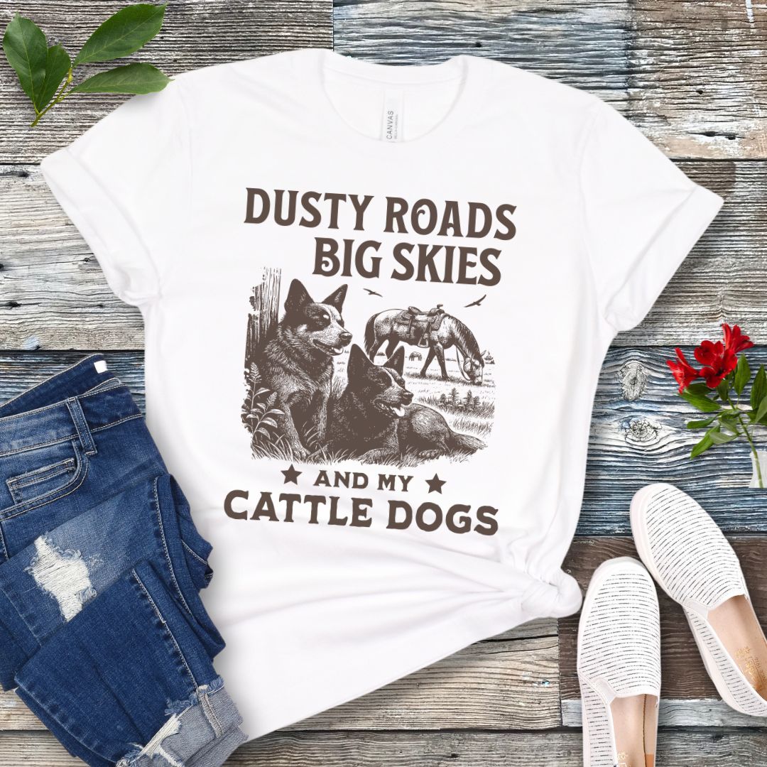 White western-aesthetic t-shirt with graphic of dogs and text 'Dusty roads, big skies, and my cattle dogs' on a wooden surface with jeans and shoes.