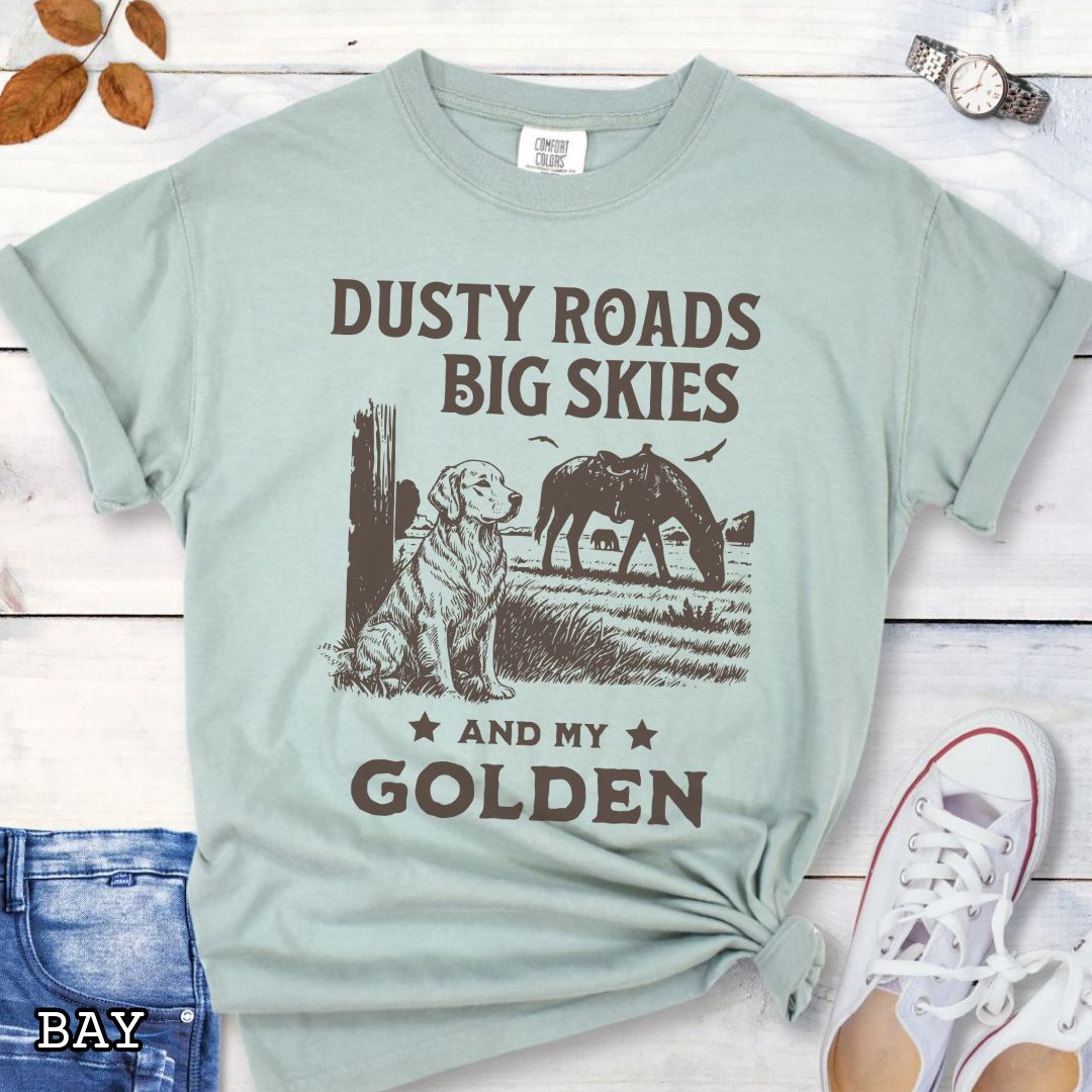 Light green Comfort Colors t-shirt featuring a Western-style design with grazing horse, rustic fence, and a Golden Retriever illustration; text reads “Dusty Roads, Big Skies, and My Golden.”