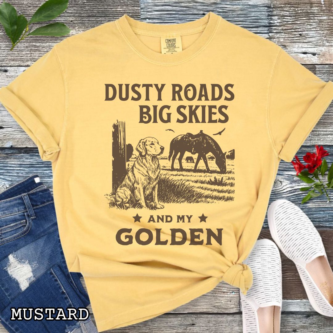Mustard-yellow Comfort Colors t-shirt featuring a Western-style design with grazing horse, rustic fence, and a Golden Retriever illustration; text reads “Dusty Roads, Big Skies, and My Golden.”