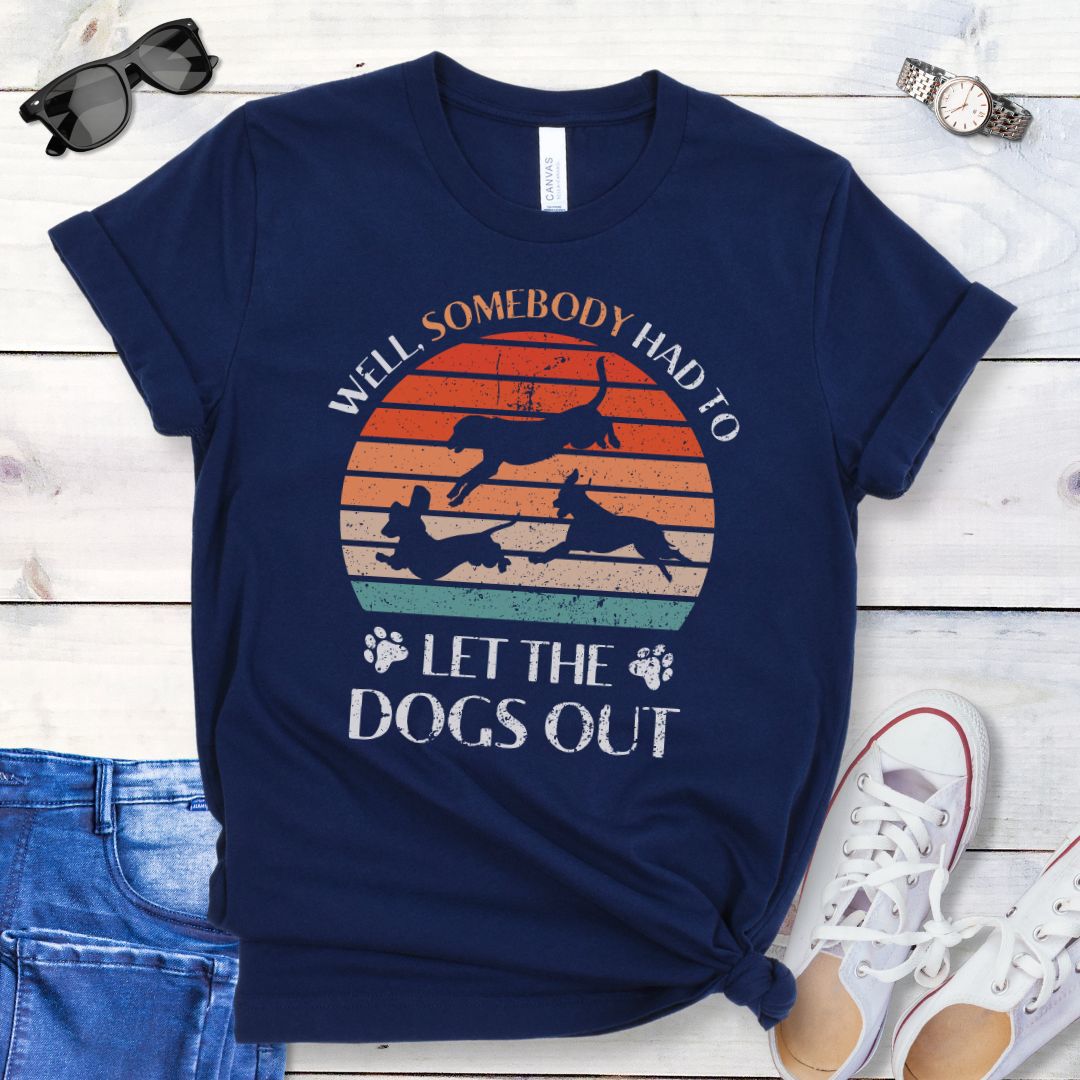A navy blue t-shirt for dog lovers with the text 'Well, Somebody Had to Let the Dogs Out' printed on the front, accompanied by a vintage sunset and an illustration of 3 dogs running.