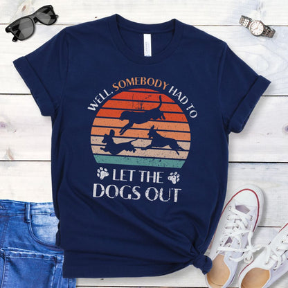 A navy blue t-shirt for dog lovers with the text 'Well, Somebody Had to Let the Dogs Out' printed on the front, accompanied by a vintage sunset and an illustration of 3 dogs running.