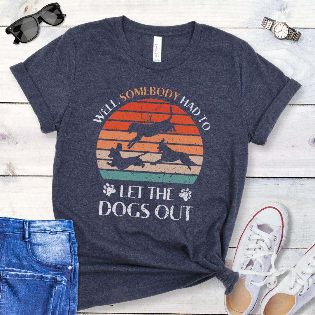 A heather navy t-shirt for dog lovers with the text 'Well, Somebody Had to Let the Dogs Out' printed on the front, accompanied by a vintage sunset and an illustration of 3 dogs running.