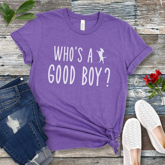 Purple unisex t-shirt with 'WHO'S A GOOD BOY? in playful font and jumping dog, presented on a wooden surface with jeans and shoes.