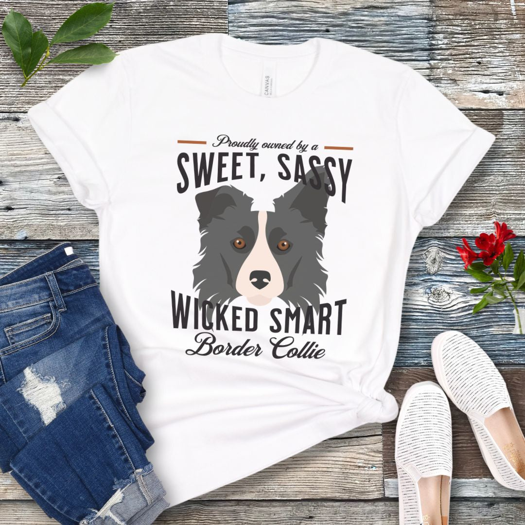 White t-shirt with border collie graphic and humorous text 'Proudly Owned by a Sweet, Sassy, Wicked Smart Border Collie', shown on a wooden surface with jeans and shoes.