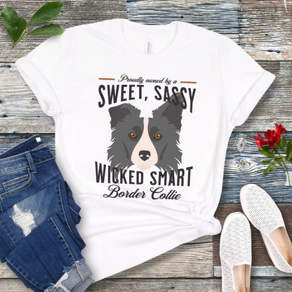 White t-shirt with border collie graphic and humorous text 'Proudly Owned by a Sweet, Sassy, Wicked Smart Border Collie', shown on a wooden surface with jeans and shoes.