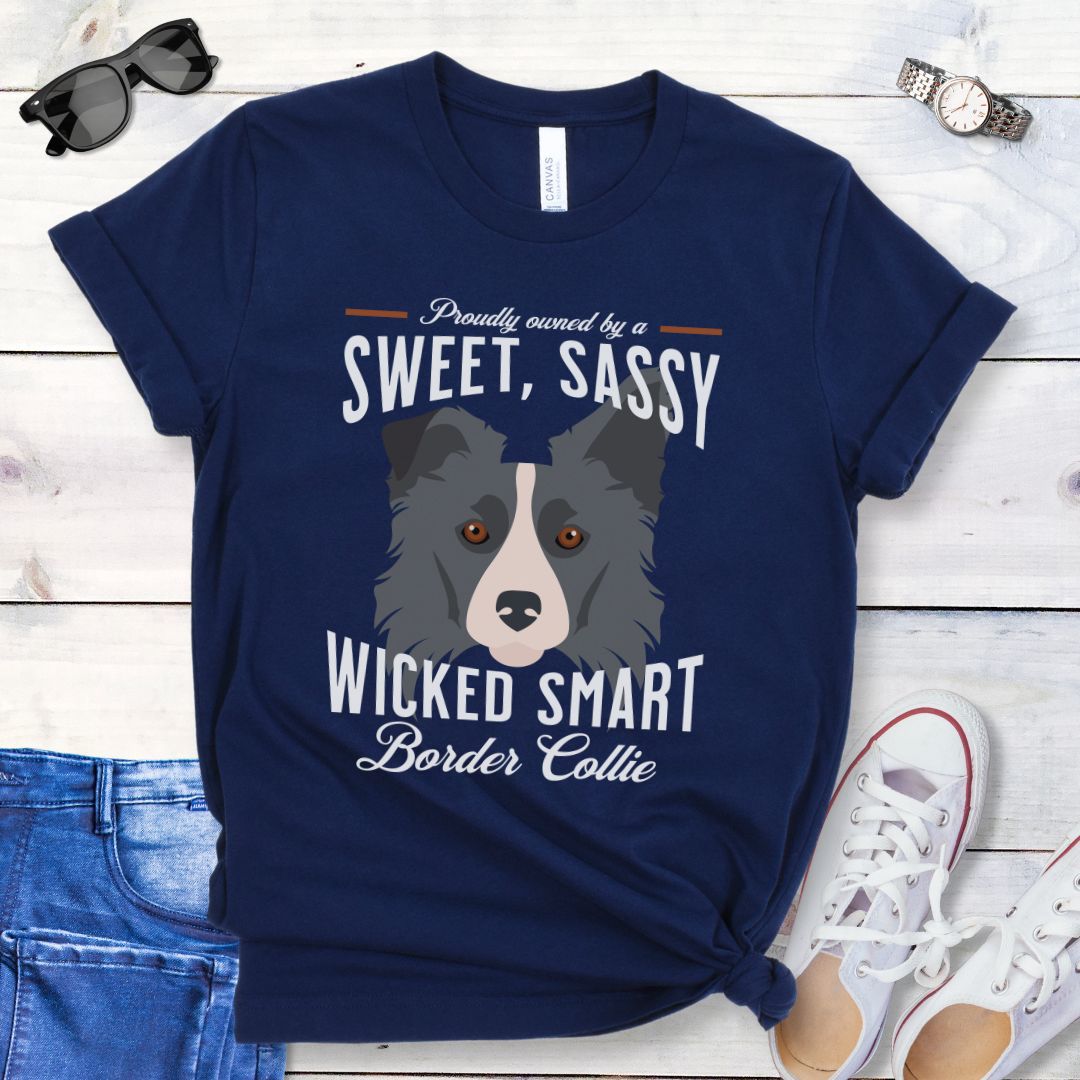 Navy t-shirt with border collie graphic and humorous text 'Proudly Owned by a Sweet, Sassy, Wicked Smart Border Collie', shown on a wooden surface with jeans and shoes.