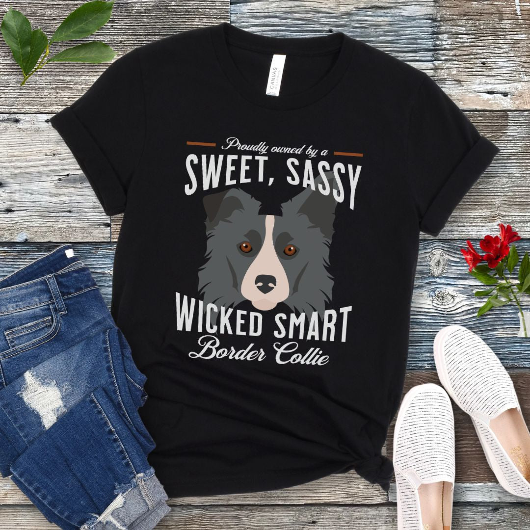 Black t-shirt with border collie graphic and humorous text 'Proudly Owned by a Sweet, Sassy, Wicked Smart Border Collie', shown on a wooden surface with jeans and shoes.