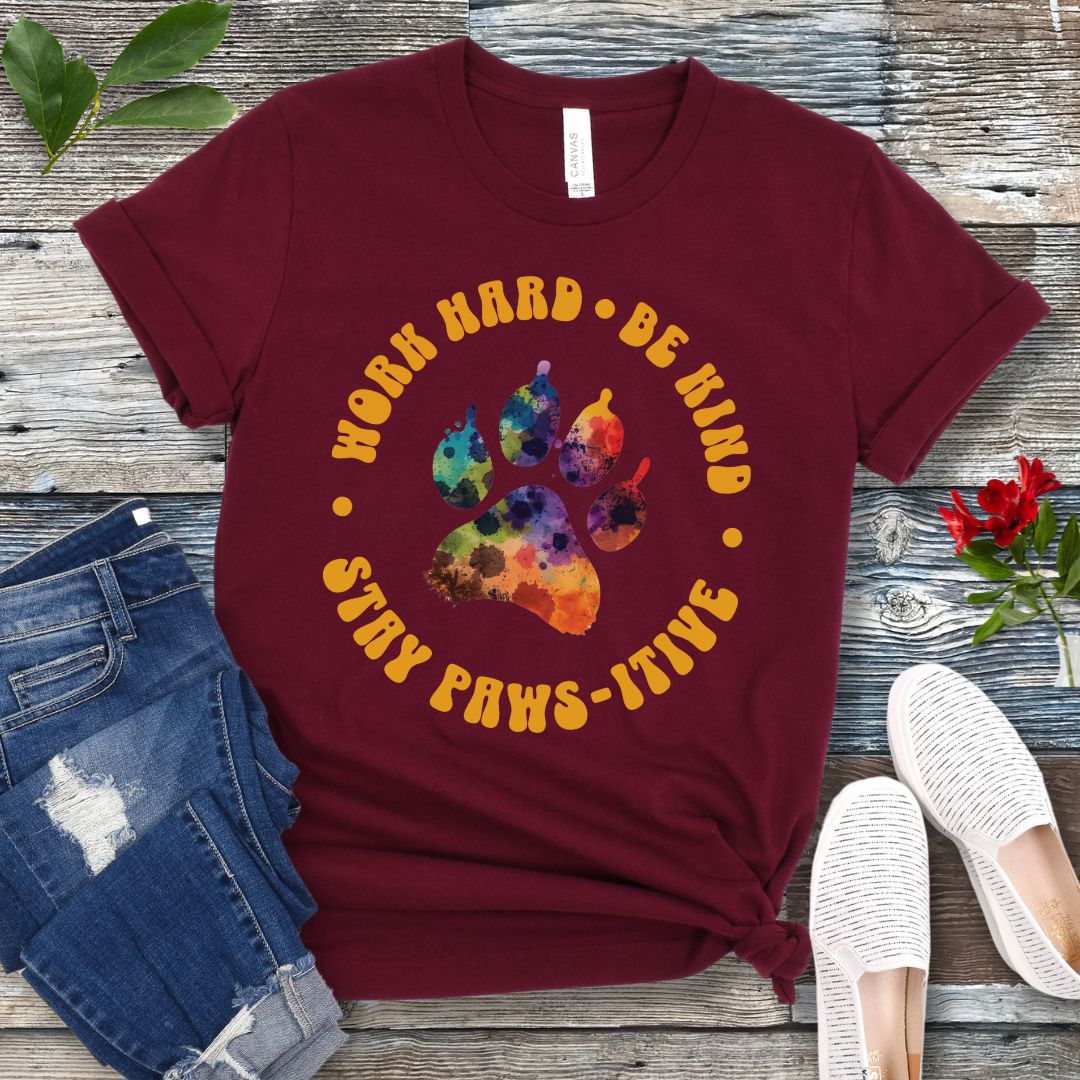 A maroon t-shirt for dog lovers with a colorful paw print design and surrounding retro-style text that reads 'WORK HARD, BE KIND, STAY PAWSITIVE'. The t-shirt is presented on a wooden surface with a pair of blue jeans and white sneakers to the side.