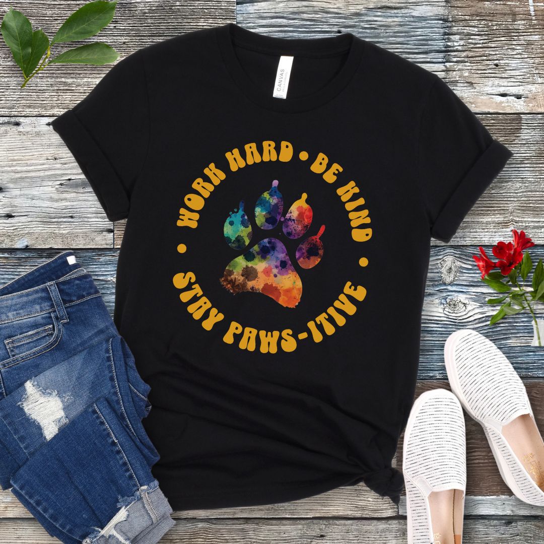 A black t-shirt for dog lovers with a colorful paw print design and surrounding retro-style text that reads 'WORK HARD, BE KIND, STAY PAWSITIVE'. The t-shirt is presented on a wooden surface with a pair of blue jeans and white sneakers to the side.