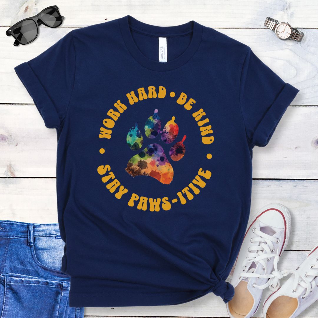 A navy t-shirt for dog lovers with a colorful paw print design and surrounding retro-style text that reads 'WORK HARD, BE KIND, STAY PAWSITIVE'. The t-shirt is presented on a wooden surface with a pair of blue jeans and white sneakers to the side.