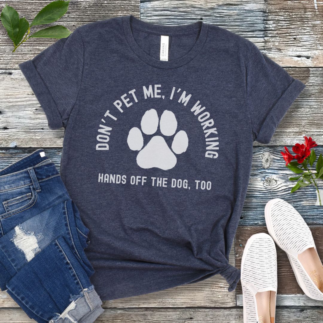 A heather navy T-shirt with the text 'DON'T PET ME, I'M WORKING. HANDS OFF THE DOG, TOO' and a paw print design, displayed on a wooden surface with a pair of jeans and white shoes in the background.